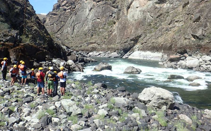 A group of students stand on a rocky shore, observing the river in front of them. The river is framed by high canyon walls. 